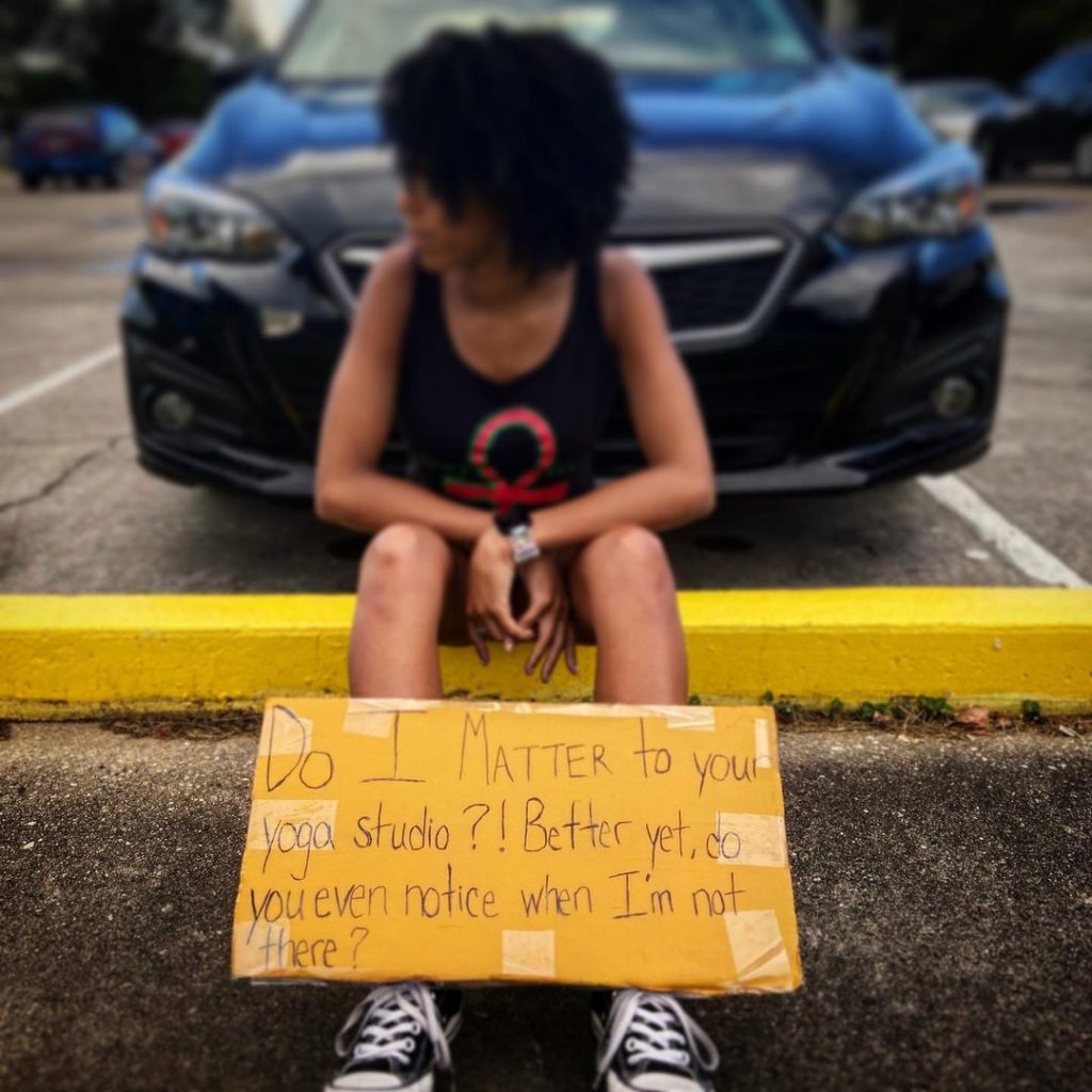 Dr. Jordan sits on a yellow parking bumper, looking off into the distance towards the left. Perched before her feet and legs is a cardboard sign reading "Do I matter to your yoga studio?! Better yet, do you even notice when I'm not there?"