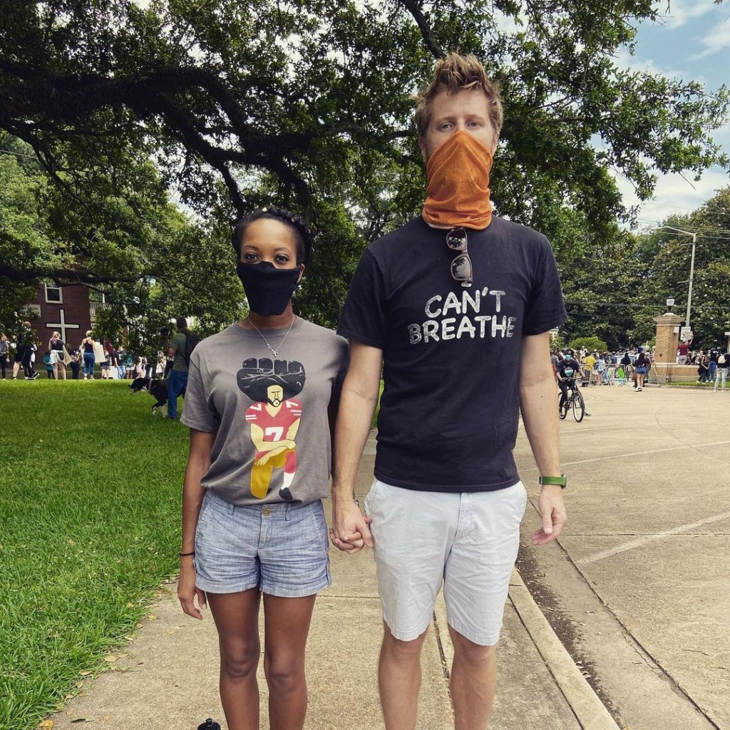 Dr. Valin Jordan and Dr. Matthew Green stand side-by-side, hands clasped. She is wearing a grey t-shirt with a cartoonized image of Colin Kaepernick kneeling and shorts. He is wearing a black t-shirt with the words "I can't Breathe" and shorts.